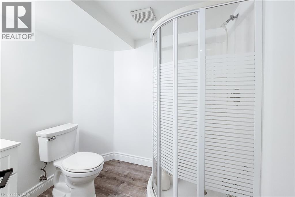 Bathroom featuring a curved shower enclosure with frosted louvered panels, a white toilet, wood-finish flooring, and white baseboards - 29 Woodside Square, Fonthill, ON - Indoor Photo Showing Bathroom