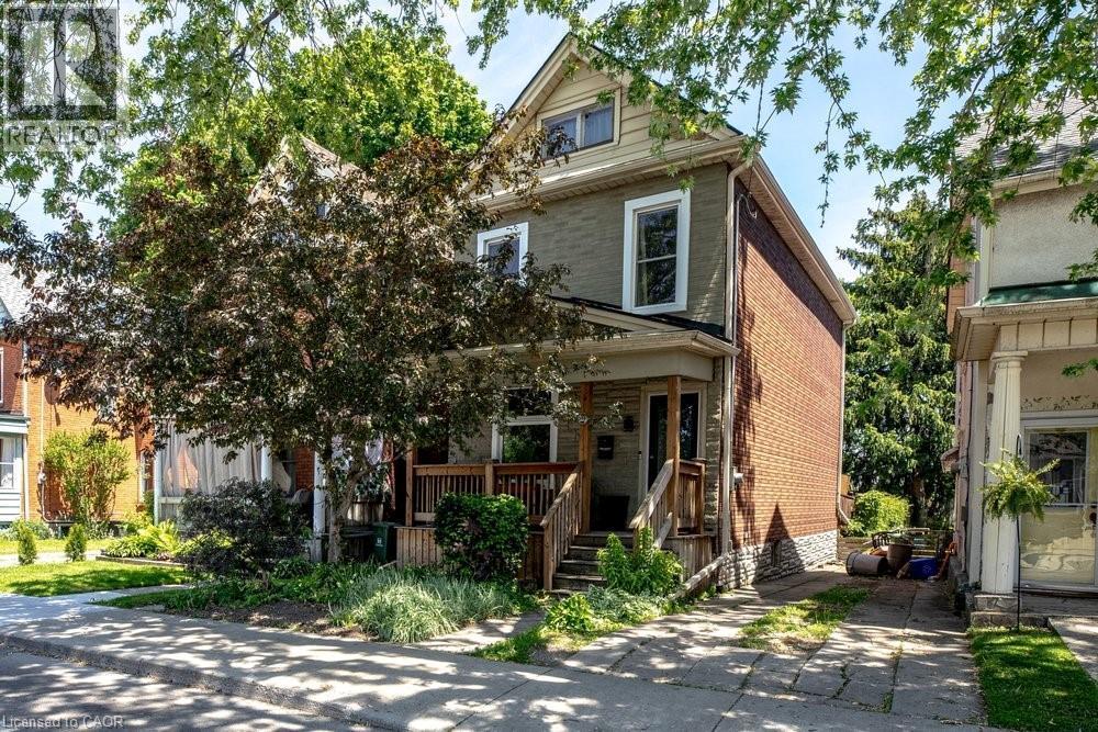 Two-story residence featuring a covered front porch with wood railings and steps, brick and siding exterior, multiple window styles, and mature landscaping - 148 Dundurn Street N, Hamilton, ON