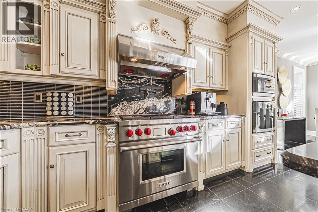 Gourmet kitchen featuring a stainless steel range with red knobs and a matching hood, custom cabinetry with decorative molding, dark patterned countertops, and integrated wall ovens - 712 Highvalley Road, Ancaster, ON