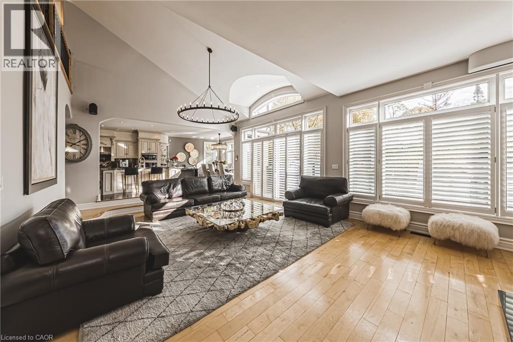 Vaulted ceiling living area featuring light wood-finish flooring, a large circular chandelier, expansive windows with plantation shutters, and an arched entryway to the kitchen - 712 Highvalley Road, Ancaster, ON