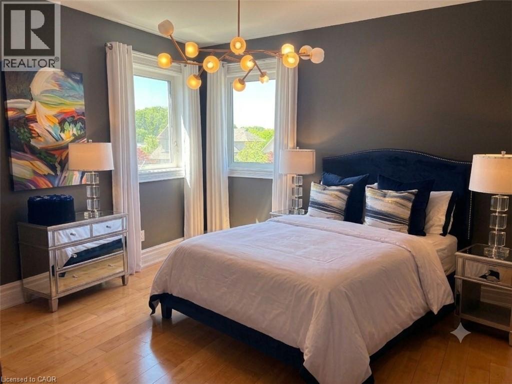 Hardwood flooring throughout this bedroom, featuring a contemporary chandelier and two large windows with white trim - 712 Highvalley Road, Ancaster, ON