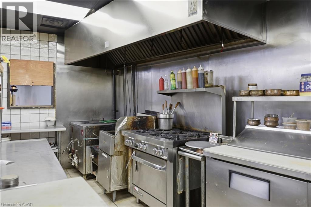 Commercial kitchen featuring a stainless steel range with multiple burners, a large stainless steel exhaust hood, stainless steel countertops, and white tiled walls - 239 York Boulevard, Hamilton, ON