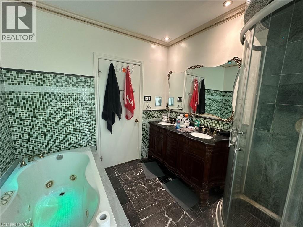 Bathroom featuring a jetted tub, a separate shower enclosure, a dual-sink vanity with a dark countertop, mosaic tile wainscoting, and patterned floor tiling - 11 Wilmont Court, Hamilton, ON