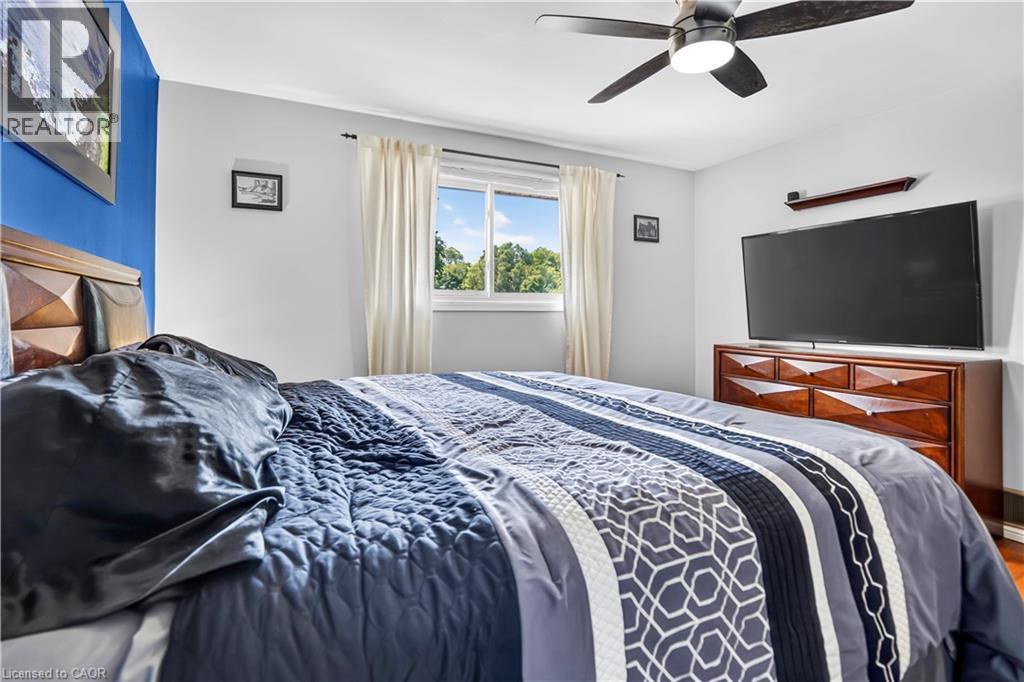 Spacious bedroom featuring light gray walls with a contrasting blue accent wall, a ceiling fan, a window with exterior tree views, and wood-finish flooring - 234 Thunderbird Drive, Cambridge, ON - Indoor Photo Showing Bedroom
