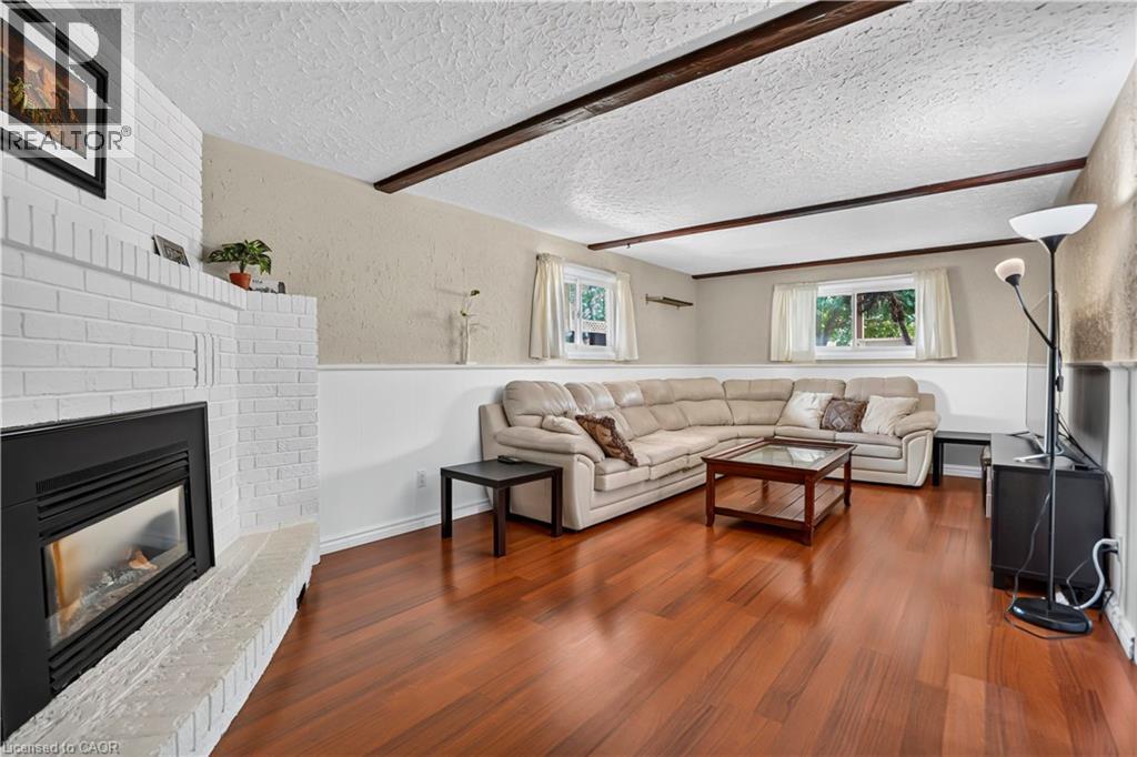 Living space featuring a white brick fireplace with a black insert, wood-finish flooring, exposed ceiling beams, and multiple windows - 234 Thunderbird Drive, Cambridge, ON - Indoor Photo Showing Living Room With Fireplace