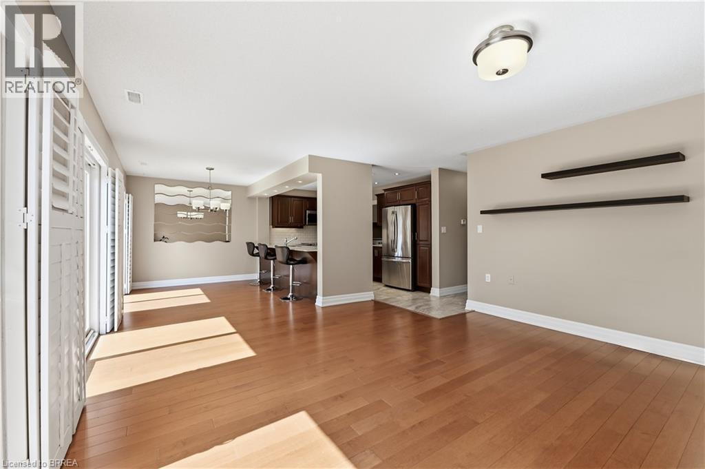 Spacious living area featuring wood-finish flooring, white plantation shutters, and a recessed kitchen with dark wood cabinetry and stainless steel appliances - 29 Archdeacon Clark Trail, Hamilton, ON