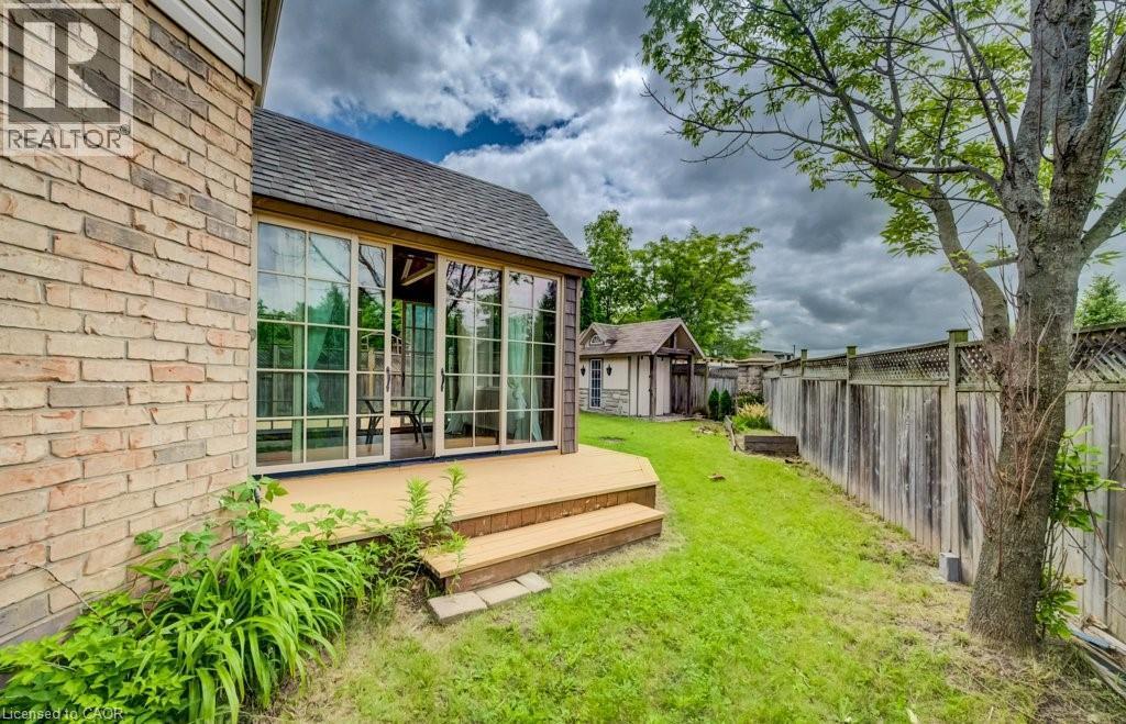 Rear exterior featuring a sunroom with multiple glass panels, an attached wooden deck with steps, a detached shed, a wooden privacy fence, and mature trees - 123 Forest Edge Trail, Kitchener, ON