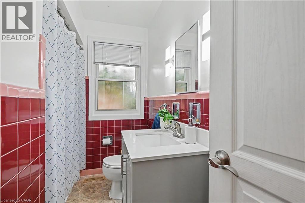 Bathroom featuring red ceramic wall tiling with pink trim, a single window with blinds, a modern vanity with a white countertop and rectangular sink, and chrome fixtures - 56 Everton Place, Hamilton, ON
