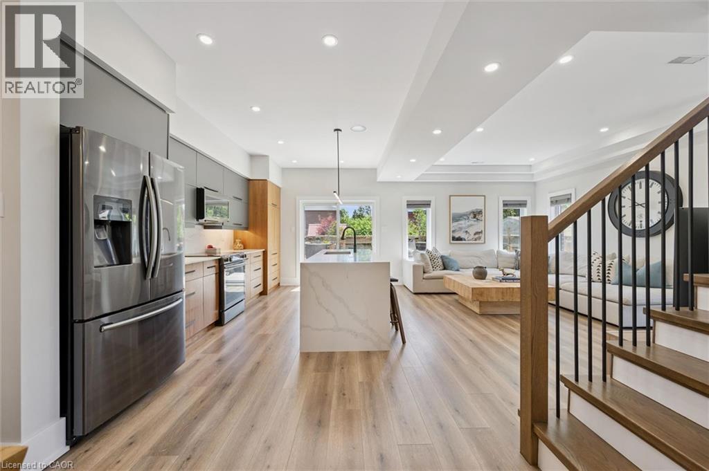 Open-concept living area featuring wood-finish flooring, a contemporary kitchen with stainless steel appliances, a waterfall island, recessed lighting, and a staircase with a wooden handrail and metal - 63 Plymouth Road, Kitchener, ON