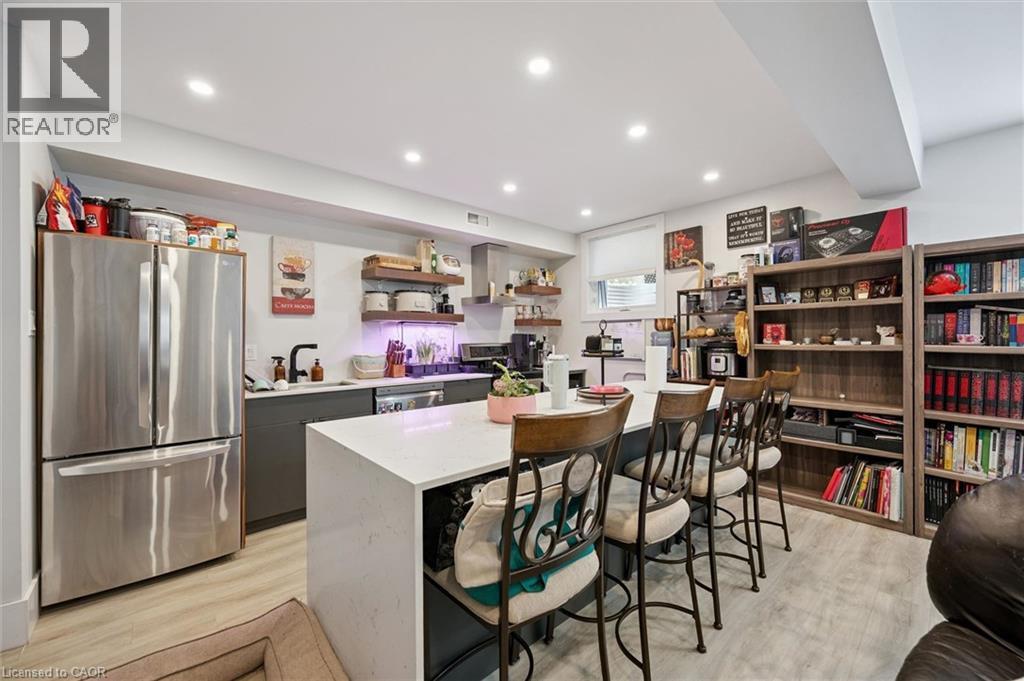 Modern kitchen area featuring a large center island with seating, recessed lighting, wood-finish flooring, stainless steel appliances, and open shelving - 63 Plymouth Road, Kitchener, ON
