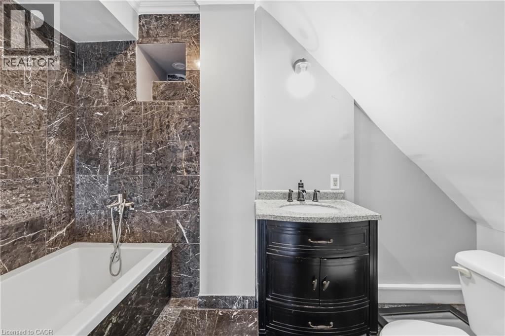 Bathroom featuring dark marbled tile walls and flooring, a white soaking tub with a wall-mounted shower fixture, a black vanity with a light speckled countertop, and an integrated sink with a dark fau - 97 Smith Road, Hamilton, ON