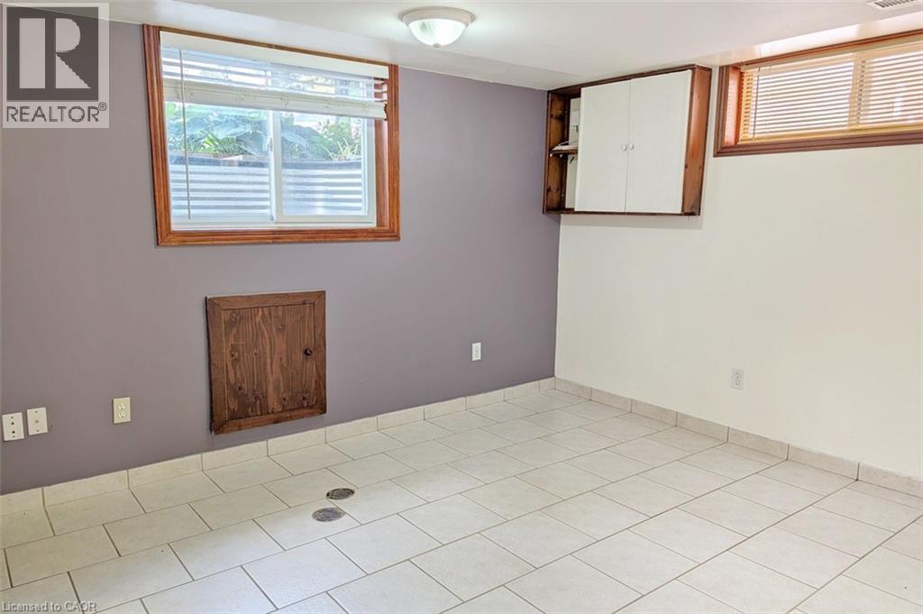 Spacious room featuring white tile flooring, two windows with wood-finish frames, a flush-mount ceiling light fixture, and a wall-mounted storage cabinet - 98 Kipling Road Unit# 2, Hamilton, ON