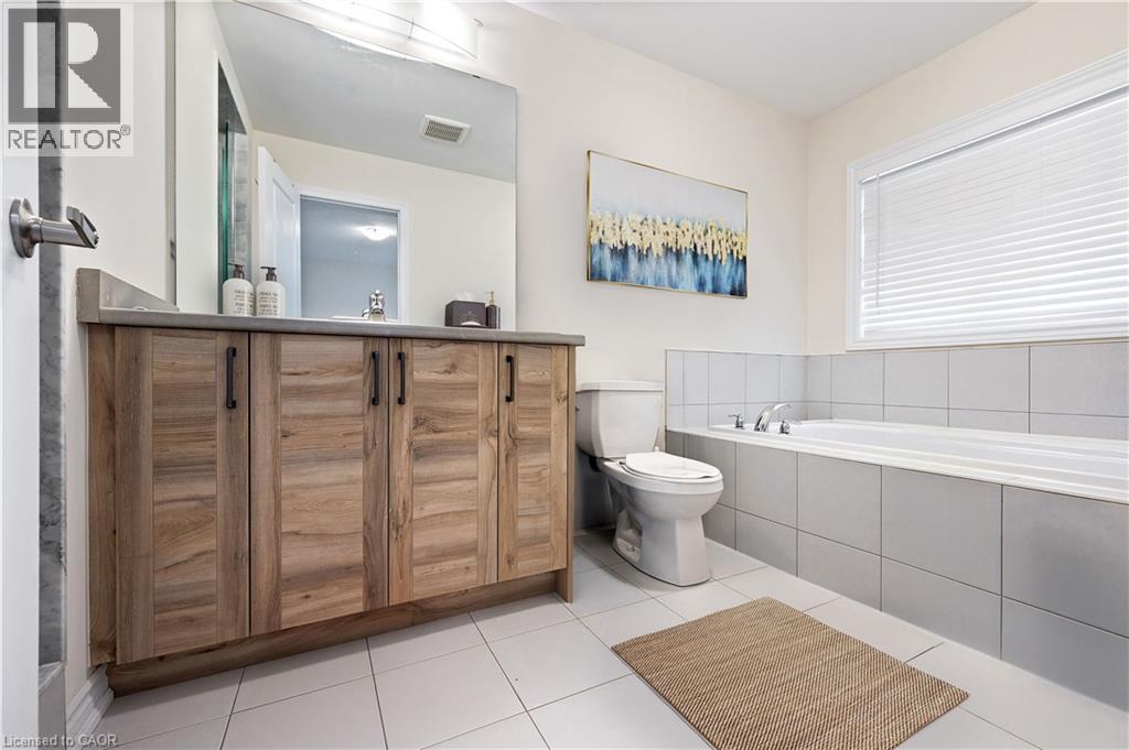 Bathroom featuring a wood-finish vanity with a light-toned countertop, a built-in bathtub with light gray tile surround, white tile flooring, and a window with blinds - 61 Vanilla Trail, Thorold, ON