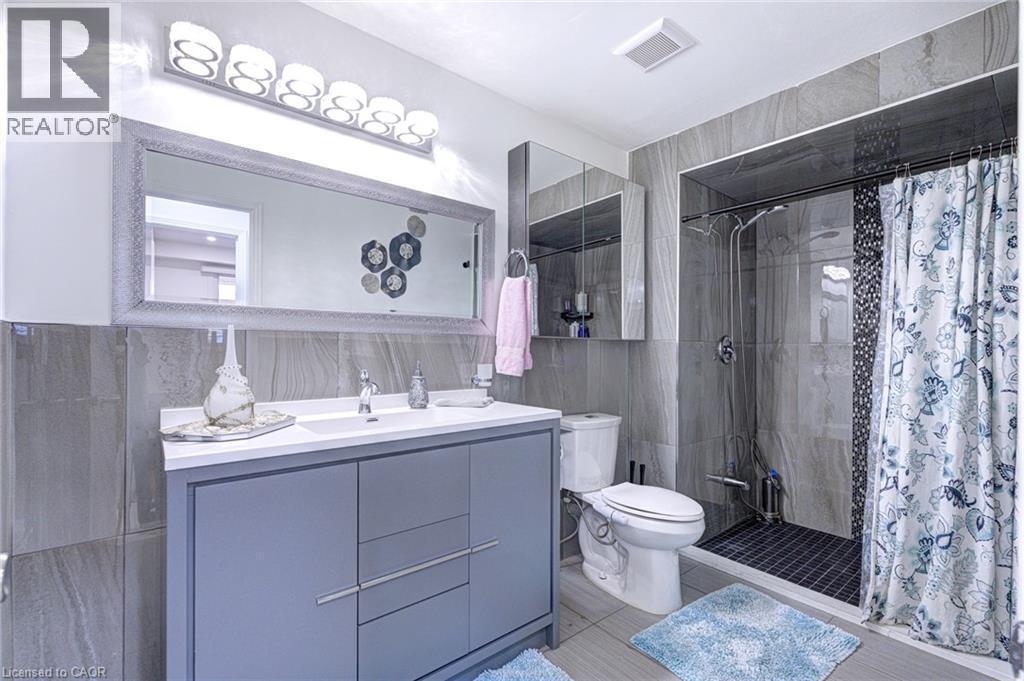 Bathroom featuring a gray vanity with an integrated sink, a large framed mirror, and a tiled shower enclosure with dark mosaic accents - 214 Merrilee Crescent, Hamilton, ON