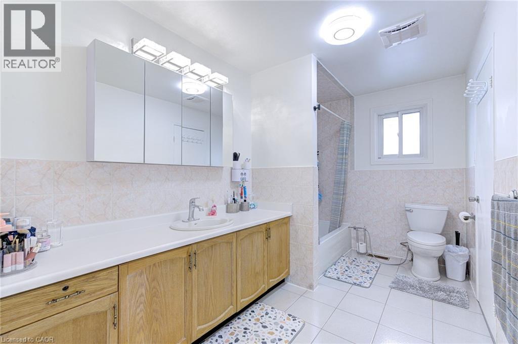 Spacious bathroom featuring a long wood-finish vanity with an integrated sink, a large mirrored medicine cabinet with vanity lighting, a full-size bathtub with an overhead shower, and white tile floor - 214 Merrilee Crescent, Hamilton, ON