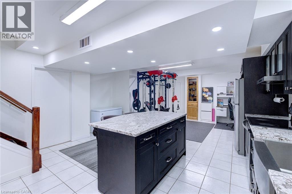 Spacious lower-level featuring a kitchen island with granite countertop and dark cabinetry, white tile flooring, recessed lighting, and a wood-finished staircase with a clear panel railing - 214 Merrilee Crescent, Hamilton, ON