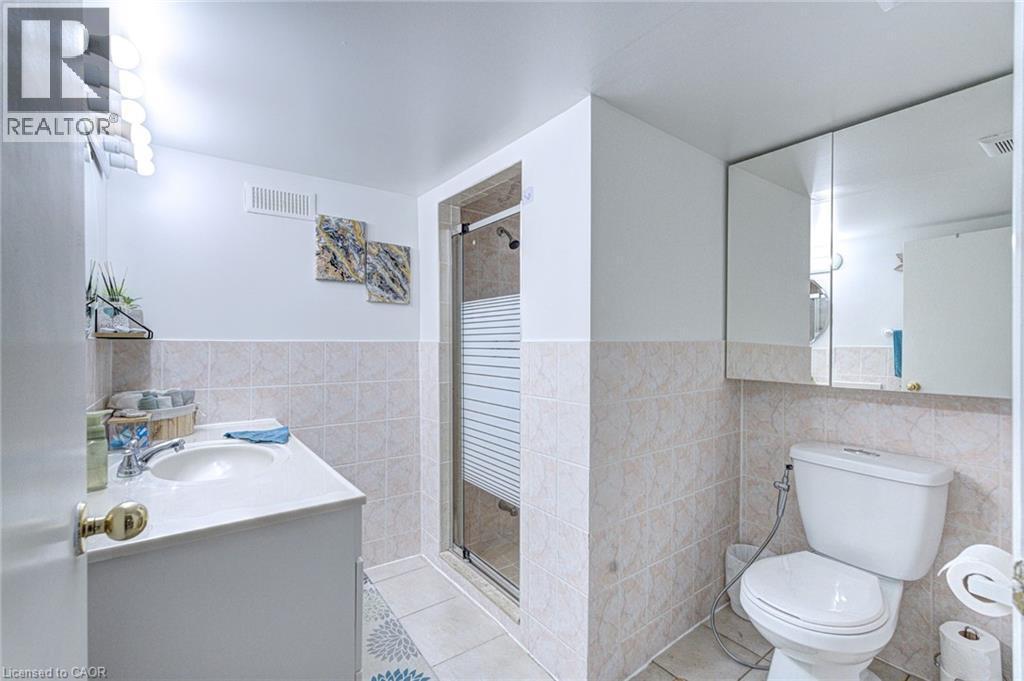 Bathroom featuring a vanity with an integrated sink, a glass-enclosed shower, a wall-mounted medicine cabinet, and light-toned floor and wall tiling - 214 Merrilee Crescent, Hamilton, ON