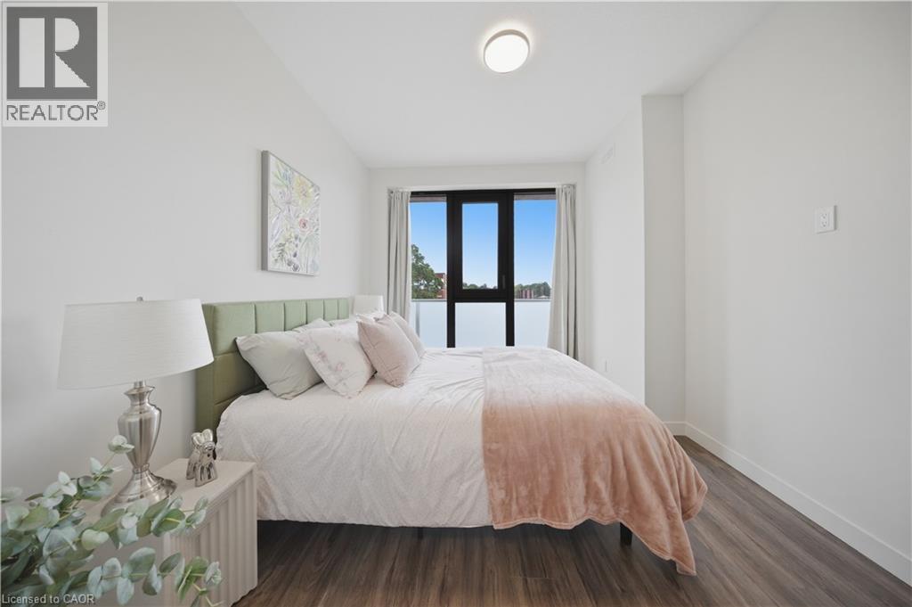 Light-filled bedroom featuring a large window with dark framing and sheer curtains, wood-finish flooring, and a modern ceiling light fixture - 1055 Weber Street E Unit# 207, Kitchener, ON