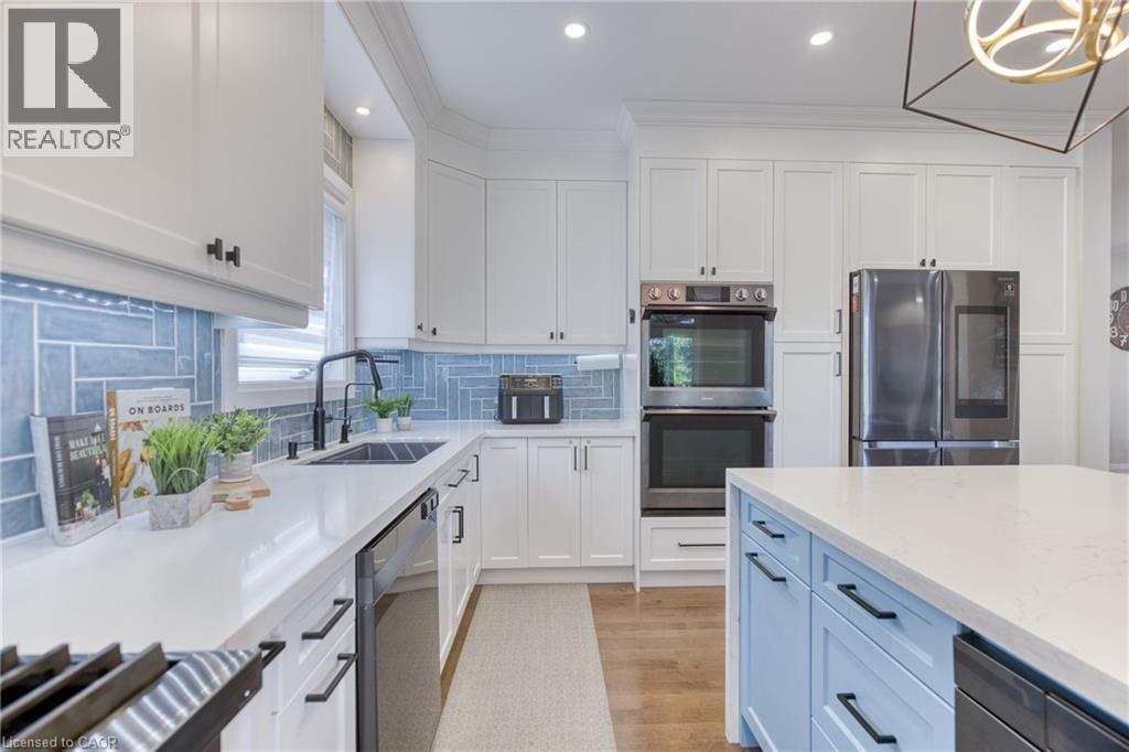 Modern kitchen featuring white cabinetry, a light blue tile backsplash, stainless steel double ovens, a stainless steel refrigerator, and a light blue kitchen island with a white countertop - 71 Ian Ormston Drive, Waterloo, ON