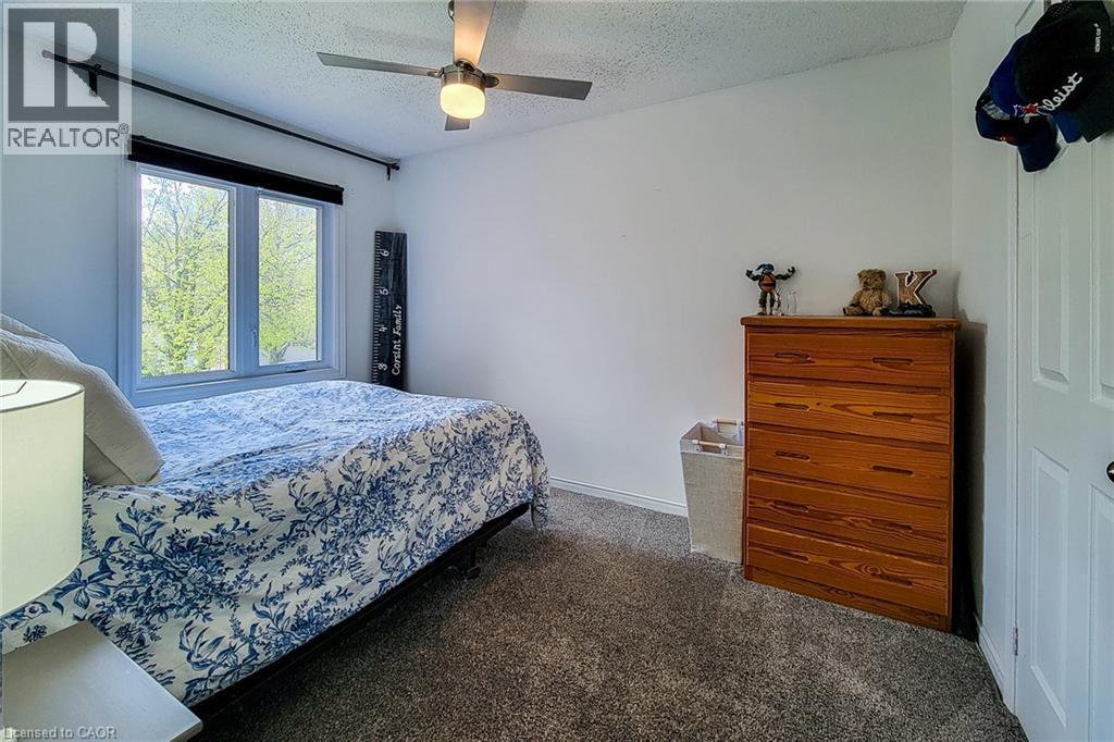 Room featuring a ceiling fan with integrated lighting, a double-pane window with a black roller shade, and neutral-toned carpeting - 7 Davidson Boulevard Unit# 14, Dundas, ON - Indoor Photo Showing Bedroom