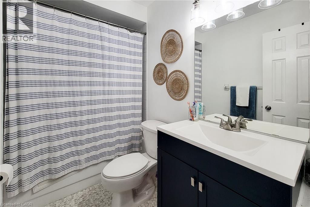 Bathroom featuring a vanity with a white countertop and dark blue cabinetry, a wall-mounted mirror, a toilet, a full shower with a tub, and light-toned flooring - 7 Davidson Boulevard Unit# 14, Dundas, ON - Indoor Photo Showing Bathroom