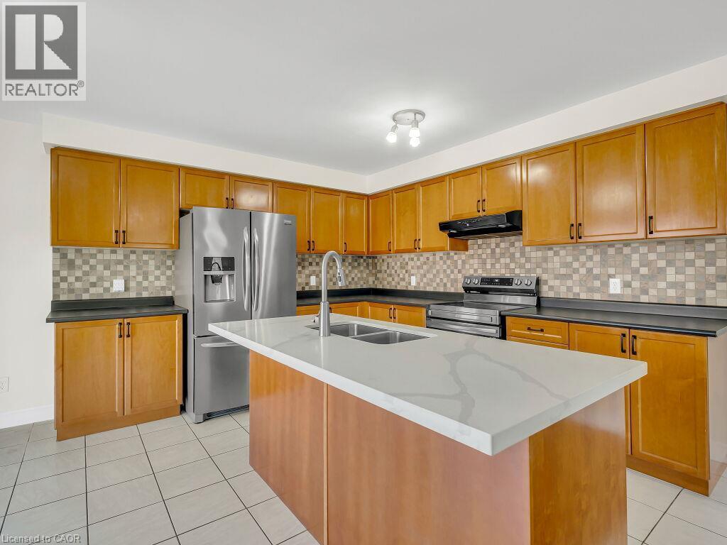 Kitchen featuring a center island with a double basin sink and a light-toned countertop, wood-finish cabinetry, stainless steel appliances, a mosaic tile backsplash, and light-toned tile flooring - 39 Baintree Way, Cambridge, ON - Indoor Photo Showing Kitchen With Stainless Steel Kitchen With Double Sink