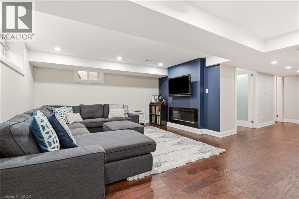 Finished lower level featuring recessed lighting, rich wood-finish flooring, a wall-mounted flat-screen television, a linear electric fireplace, and a partial glass-paneled door - 715 Drury Lane, Burlington, ON - Indoor With Fireplace
