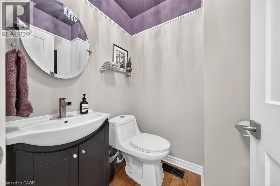 Powder room featuring a curved vanity with a white basin, a wall-mounted round mirror, and wood-finish flooring - 30 Carriagegate Drive, Hamilton, ON - Indoor Photo Showing Bathroom