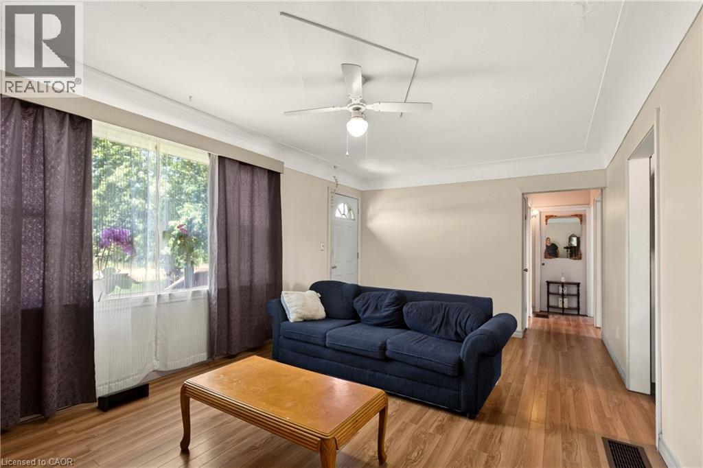 Living area featuring wood-finish flooring, a ceiling fan with integrated lighting, and a large window with sheer and blackout curtain options - 958 Highland Road E, Hamilton, ON