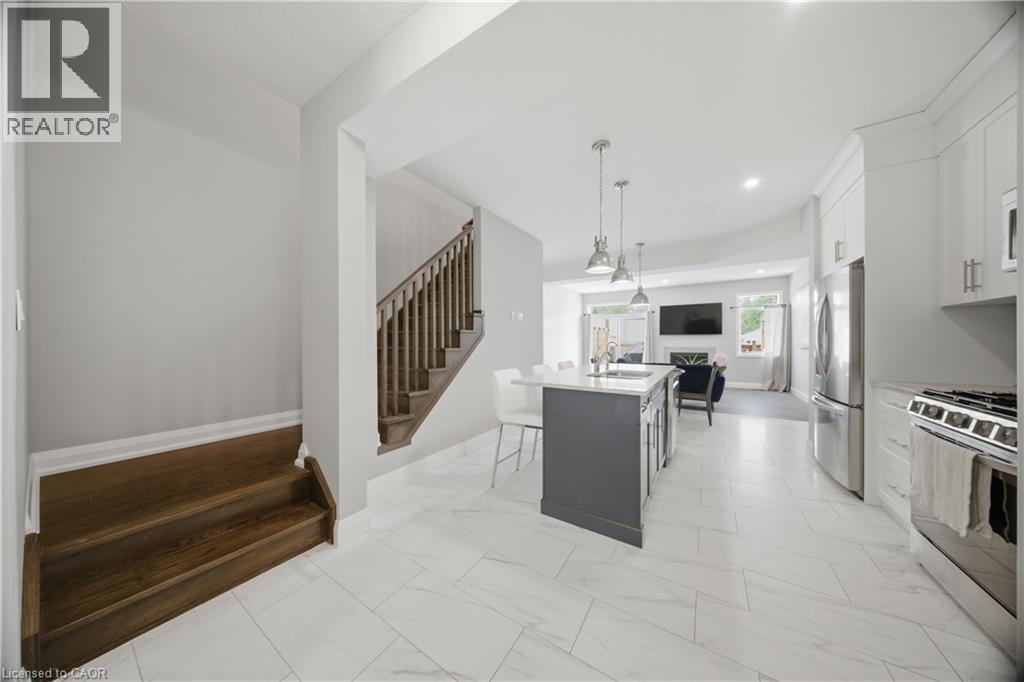 Open-concept kitchen and living area featuring white tile flooring, a dark-toned kitchen island with pendant lighting, stainless steel appliances, white cabinetry, and a wood-finish staircase - 107 Stephenson Way, Palmerston, ON - Indoor Photo Showing Kitchen With Stainless Steel Kitchen