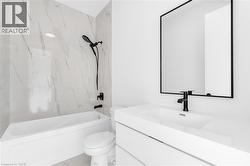 Bathroom featuring a white bathtub with a matte black shower head and faucet, marble-look tiled shower surround, white toilet, and a vanity with an integrated sink and matte black faucet -