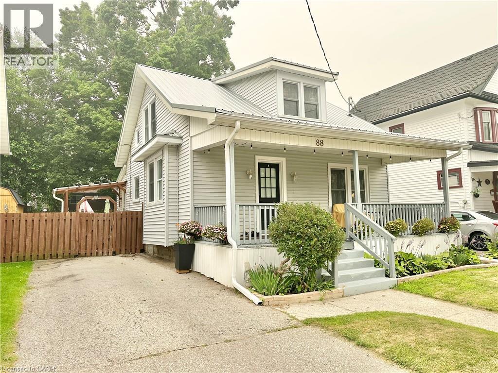 Charming light-colored siding home featuring a covered front porch with railings, a metal roof, and a black front door - 88 Brook Street, Simcoe, ON - Outdoor With Deck Patio Veranda