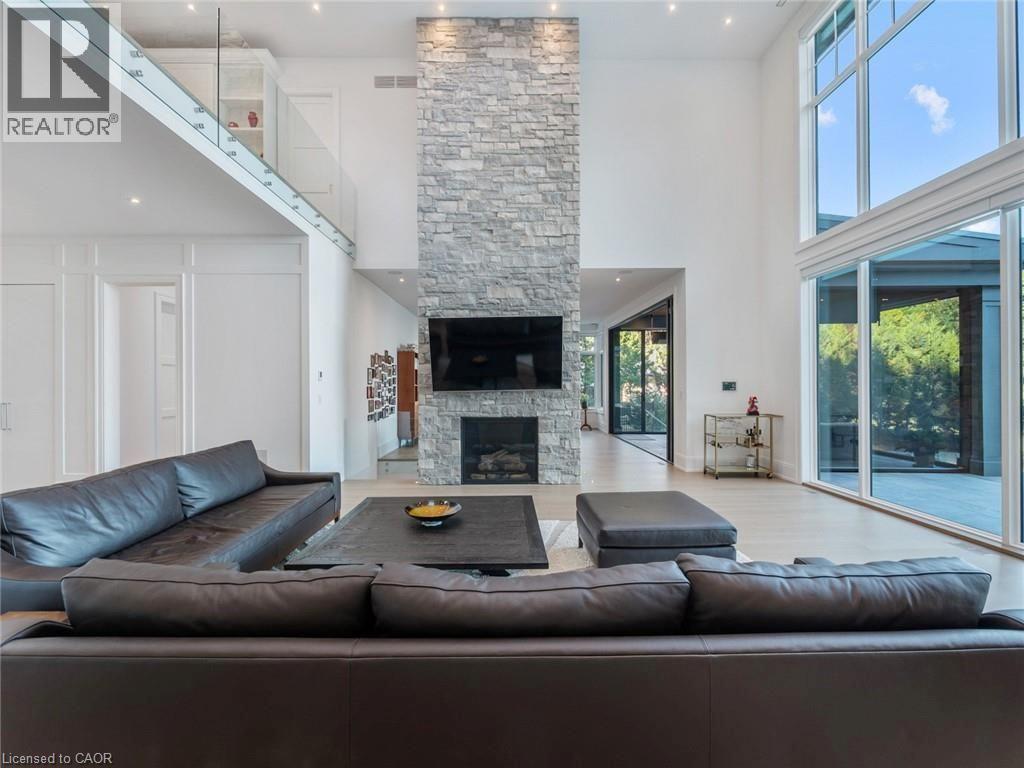 Double-height living area featuring a floor-to-ceiling stacked stone fireplace, large format windows, light wood-finish flooring, and a second-story glass railing balcony - 140 Secord Lane, Burlington, ON - Indoor Photo Showing Living Room With Fireplace