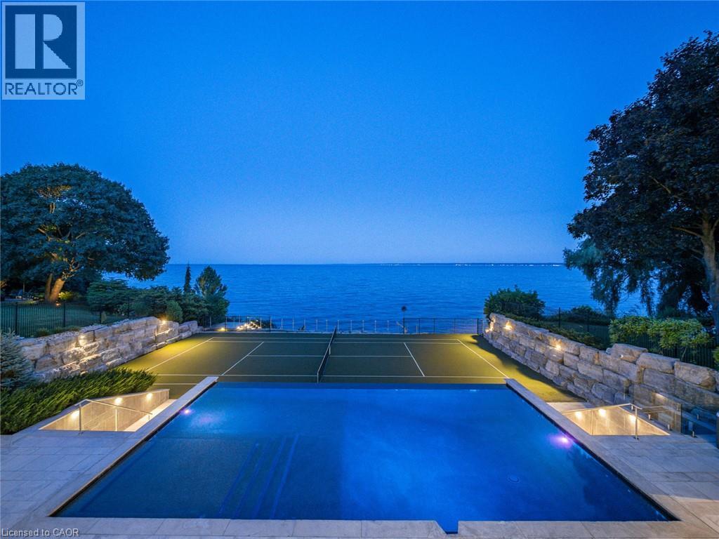 Expansive waterside grounds featuring an illuminated tennis court, a covered swimming pool, and stone retaining walls with accent lighting - 140 Secord Lane, Burlington, ON - Outdoor With Body Of Water With In Ground Pool