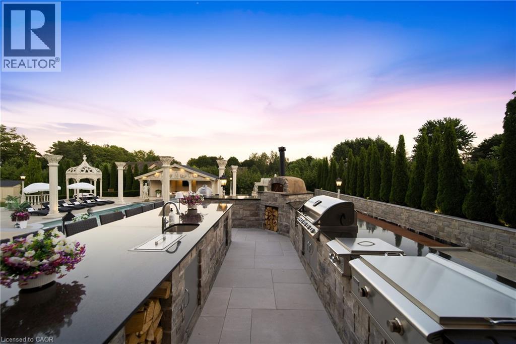 Outdoor kitchen featuring extensive countertop space, an integrated sink, built-in stainless steel appliances, a pizza oven, and stacked stone finishes - 7009 Wellington Rd 34, Puslinch, ON - Outdoor