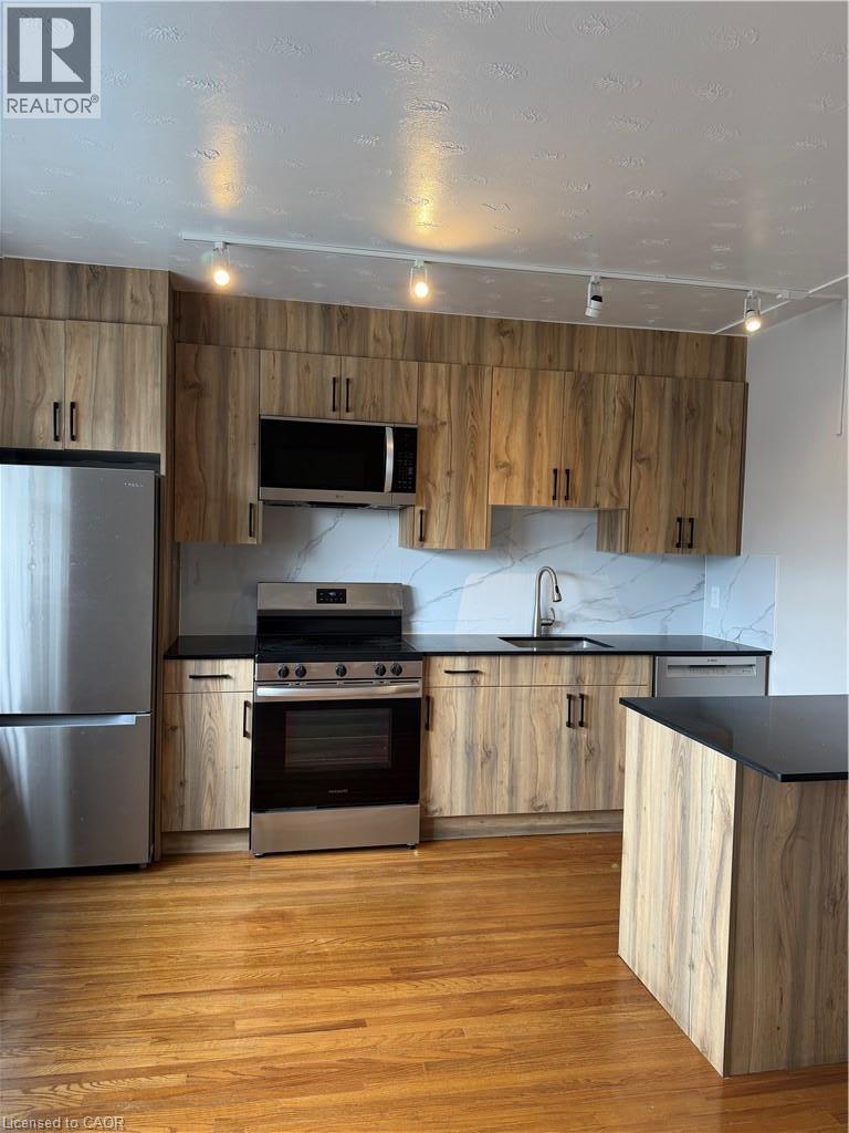 Modern kitchen featuring wood-finish cabinetry, stainless steel appliances, black countertops, and a white tile backsplash - 237 Mohawk Road E Unit# 16, Hamilton, ON - Indoor Photo Showing Kitchen