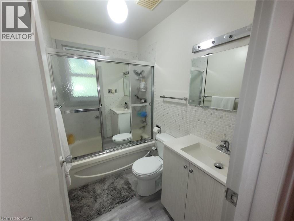 Bathroom featuring a full tub with sliding glass enclosure, mosaic tile wainscoting, vanity with integrated sink, and wood-finish flooring - 879 West 5Th Street, Hamilton, ON - Indoor Photo Showing Bathroom