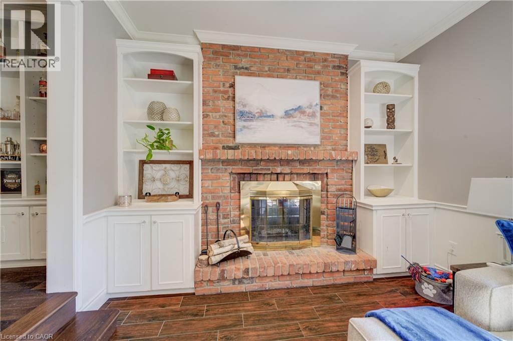 Brick fireplace with brass firebox and hearth, flanked by built-in white cabinetry and shelving, wood-finish flooring, and crown molding - 41 Mcniven Road, Ancaster, ON - Indoor With Fireplace