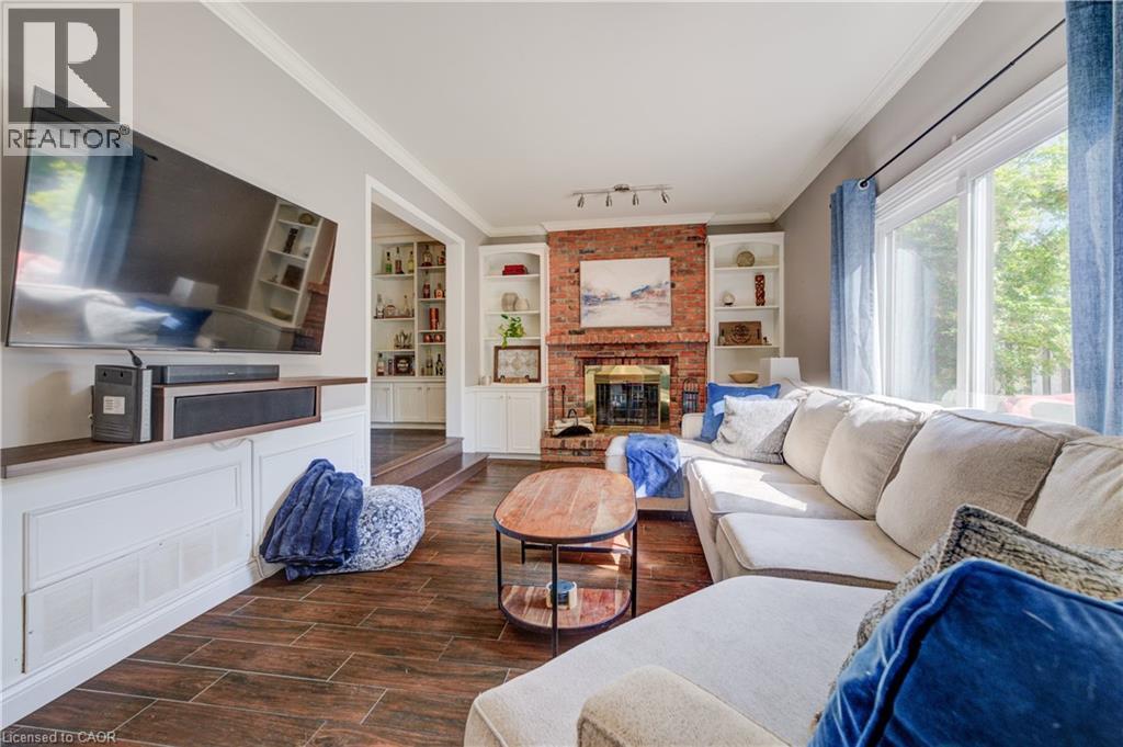 Living area featuring wood-finish flooring, a brick fireplace with a brass firebox, built-in shelving, white baseboards, and a large window - 41 Mcniven Road, Ancaster, ON - Indoor Photo Showing Living Room With Fireplace