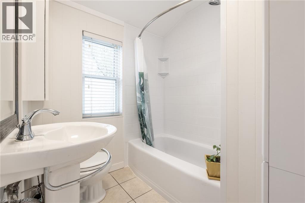 Bathroom featuring a white pedestal sink with chrome faucet, a white bathtub with a tiled surround, and a window with horizontal blinds - 851 Beach Boulevard, Hamilton, ON - Indoor Photo Showing Bathroom