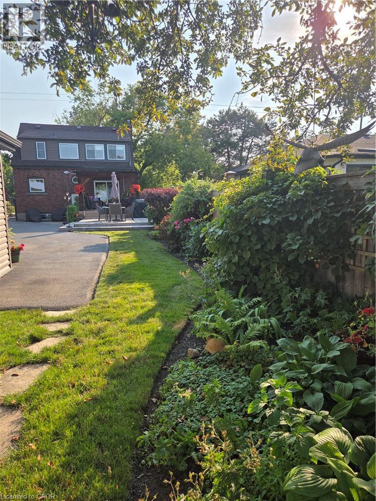 Expansive backyard featuring a manicured lawn, established garden beds with diverse foliage, a paved patio area, and a red brick exterior with a second-story dormer - 851 Beach Boulevard, Hamilton, ON - Outdoor With Deck Patio Veranda