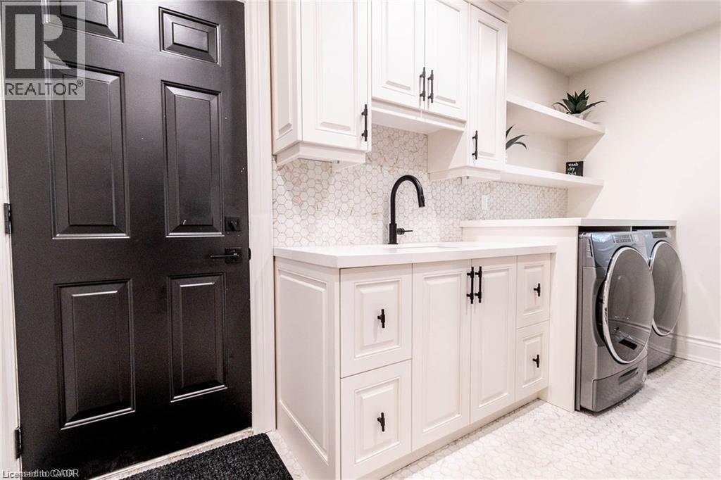 Utility area featuring a paneled black door, white shaker cabinetry with black hardware, hexagon tile backsplash, and a built-in countertop above laundry appliances - 76 Adriatic Boulevard, Hamilton, ON - Indoor Photo Showing Laundry Room