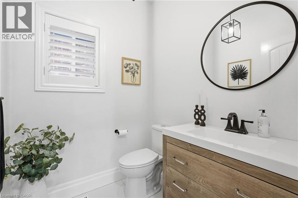 Powder room featuring a single-sink wood-finish vanity with a white countertop and matte black faucet, a round black-framed mirror, and a window with white louvered shutters - 47 Winners Way, Binbrook, ON - Indoor Photo Showing Bathroom