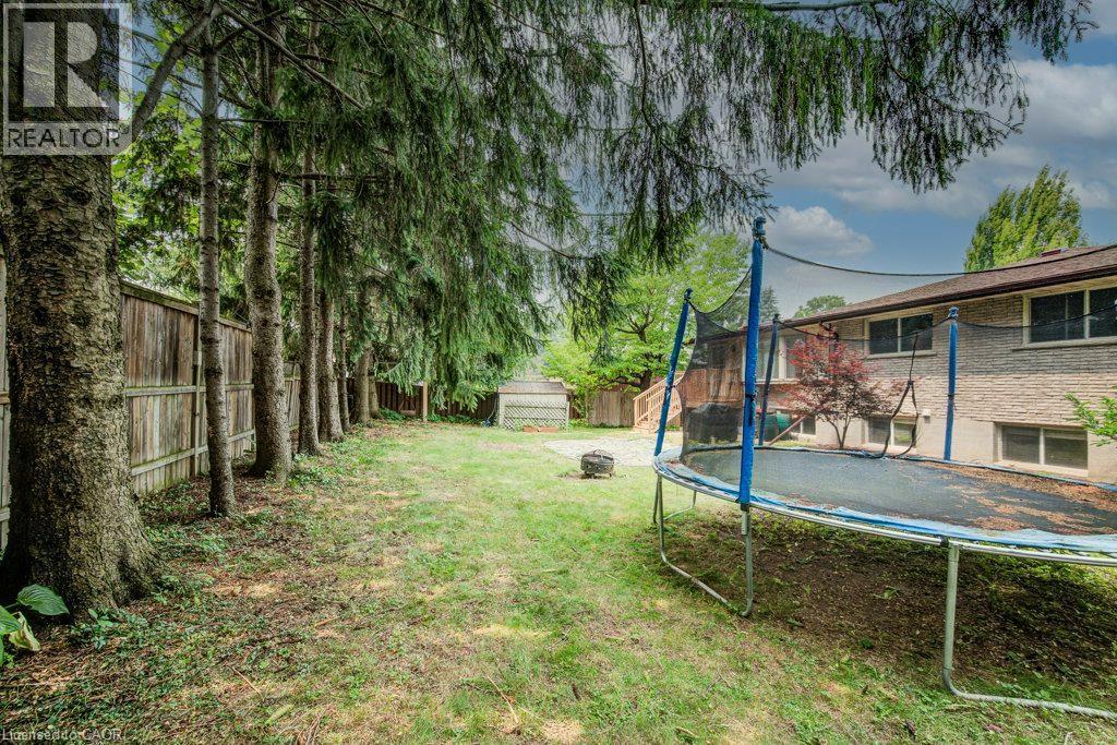 Expansive rear yard featuring mature evergreen trees, a wooden privacy fence, natural grass coverage, a fire pit, and a brick exterior dwelling with multiple windows and a brown shingle roof - 109 White Oak Place, Waterloo, ON - Outdoor