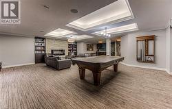 Spacious recreation room featuring patterned carpet flooring, a stone-clad fireplace, built-in bookshelves, tray ceilings with integrated lighting, and a prominent billiards table -