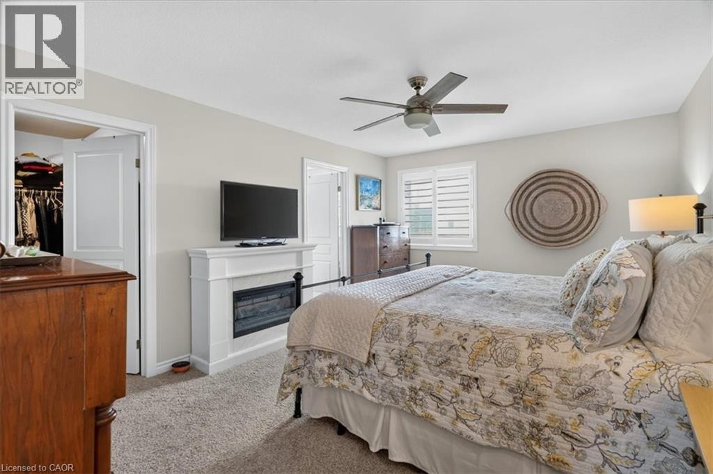 Carpeted bedroom featuring a ceiling fan, window with white louvered shutters, and a white fireplace surround with a dark insert - 87 Boyd Boulevard, Thamesford, ON - Indoor Photo Showing Bedroom