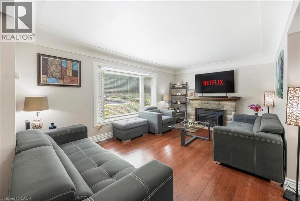 Spacious living area featuring wood-finish flooring, a stone-clad fireplace with a dark metal insert, and a large bay window - 1063 Joan Drive, Burlington, ON - Indoor Photo Showing Living Room With Fireplace