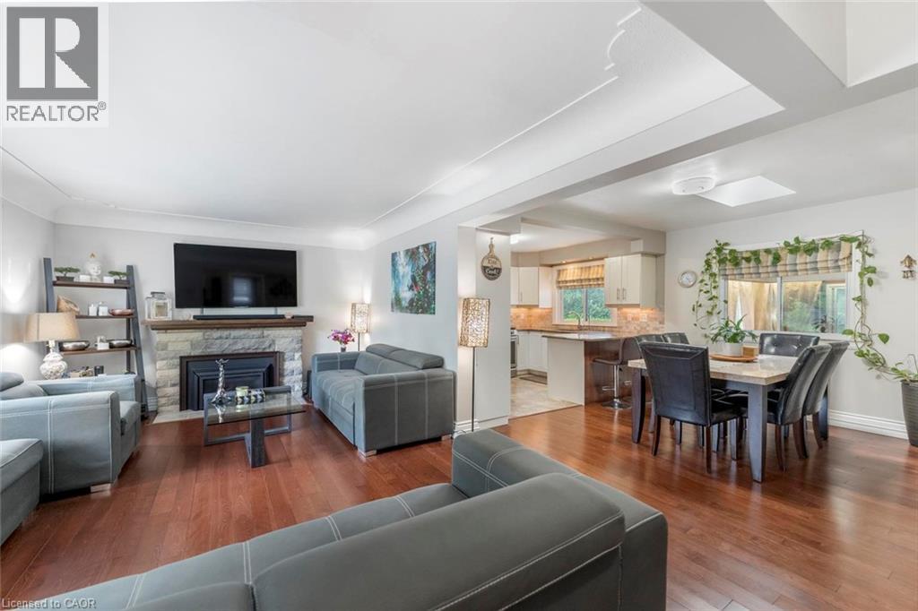 Open-concept living space featuring wood-finish flooring, a stone-surround fireplace with a wood mantel, a dining area with a rectangular table, and a partial kitchen with an island - 1063 Joan Drive, Burlington, ON - Indoor Photo Showing Living Room With Fireplace