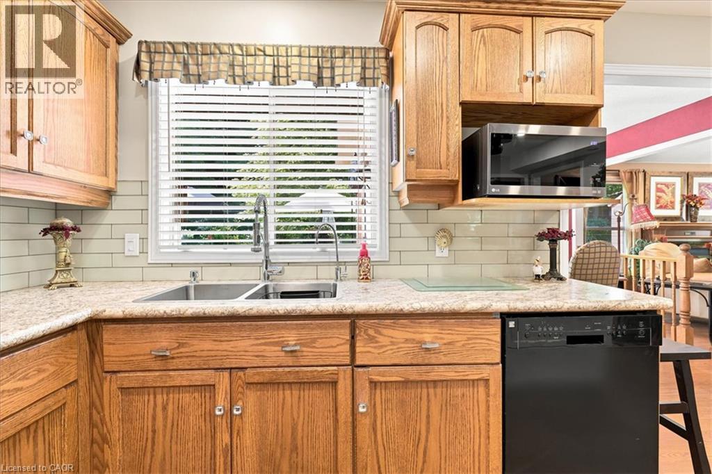 Kitchen workstation featuring wood cabinetry, light-toned countertops, a stainless steel double basin sink with a gooseneck faucet, a light subway tile backsplash, and an over-the-range microwave - 39 Everwood Run, Kitchener, ON - Indoor Photo Showing Kitchen With Double Sink