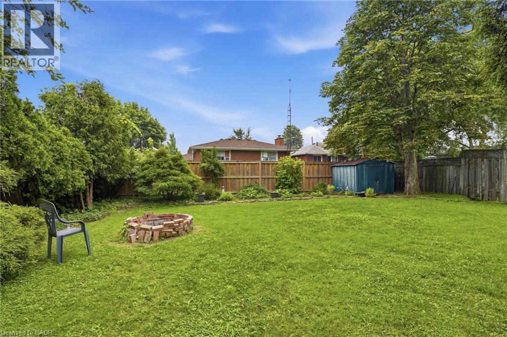 Expansive green lawn with a brick fire pit, mature trees, and a wooden privacy fence - 834 Mohawk Road E, Hamilton, ON - Outdoor With Backyard